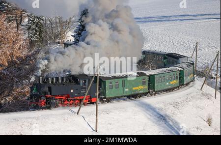 Cranzahl, Germany. 13th Dec, 2022. The Fichtelberg Railway makes its way through the snowy winter forest in the Ore Mountains with plenty of smoke and clouds of smoke. The narrow-gauge railroad is struggling with increased coal prices. The Sächsisches Dampfeisenbahngesellschaft mbH (SDG) needs around 800 tons of coal per year for the Fichtelberg, Lößnitzgrund and Weißeritztalbahn. To save money, it therefore now only operates one steam locomotive on the line. The other railroads are also thinning out operations at off-peak times. Credit: Jan Woitas/dpa/Alamy Live News Stock Photo