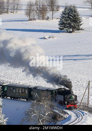 Cranzahl, Germany. 13th Dec, 2022. The Fichtelberg Railway makes its way through the snowy winter forest in the Ore Mountains with plenty of smoke and clouds of smoke. The narrow-gauge railroad is struggling with increased coal prices. The Sächsisches Dampfeisenbahngesellschaft mbH (SDG) needs around 800 tons of coal per year for the Fichtelberg, Lößnitzgrund and Weißeritztalbahn. To save money, it therefore now only operates one steam locomotive on the line. The other railroads are also thinning out operations at off-peak times. Credit: Jan Woitas/dpa/Alamy Live News Stock Photo