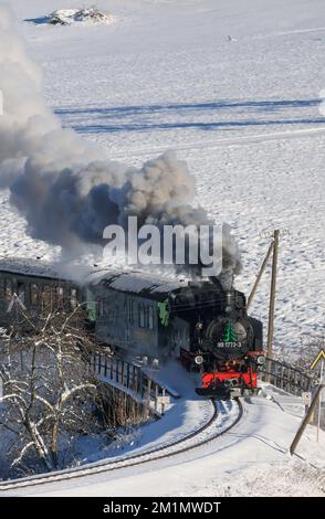 Cranzahl, Germany. 13th Dec, 2022. The Fichtelberg Railway makes its way through the snowy winter forest in the Ore Mountains with plenty of smoke and clouds of smoke. The narrow-gauge railroad is struggling with increased coal prices. The Sächsisches Dampfeisenbahngesellschaft mbH (SDG) needs around 800 tons of coal per year for the Fichtelberg, Lößnitzgrund and Weißeritztalbahn. To save money, it therefore now only operates one steam locomotive on the line. The other railroads are also thinning out operations at off-peak times. Credit: Jan Woitas/dpa/Alamy Live News Stock Photo