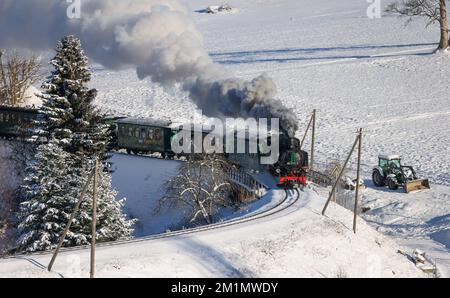 Cranzahl, Germany. 13th Dec, 2022. The Fichtelberg Railway makes its way through the snowy winter forest in the Ore Mountains with plenty of smoke and clouds of smoke. The narrow-gauge railroad is struggling with increased coal prices. The Sächsisches Dampfeisenbahngesellschaft mbH (SDG) needs around 800 tons of coal per year for the Fichtelberg, Lößnitzgrund and Weißeritztalbahn. To save money, it therefore now only operates one steam locomotive on the line. The other railroads are also thinning out operations at off-peak times. Credit: Jan Woitas/dpa/Alamy Live News Stock Photo