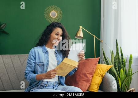 Happy woman at home received nice notification letter reading and happy smiling, Hispanic woman at home with holding message envelope sitting on sofa in living room. Stock Photo