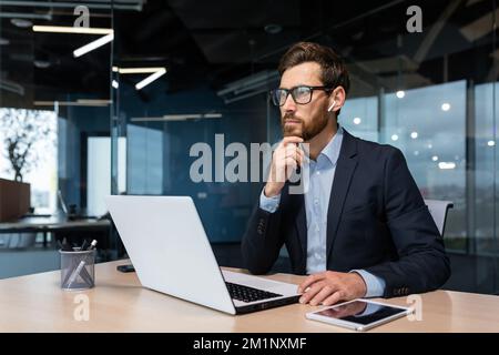 Thoughtful and serious young man businessman, freelancer, designer, architect works in the office at a laptop with headphones. Stock Photo