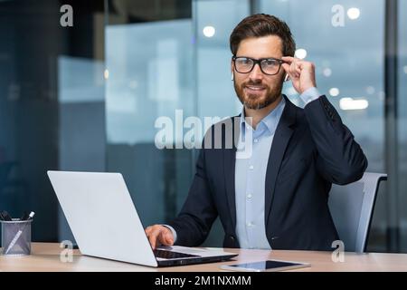 Portrait of a successful young businessman, lawyer, legal defender. Confidently looks into the camera, holds glasses, laughs, sits in the office at the table, works at the laptop. Stock Photo