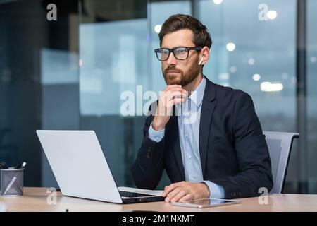 Portrait of a young serious male businessman in the office at the table in headphones and with a laptop. Looks to the side. Stock Photo