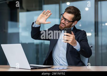 Young handsome man sitting in a suit in the office at the table and listening to music from the phone in headphones. Dances along, relaxed, waves his hands. Stock Photo