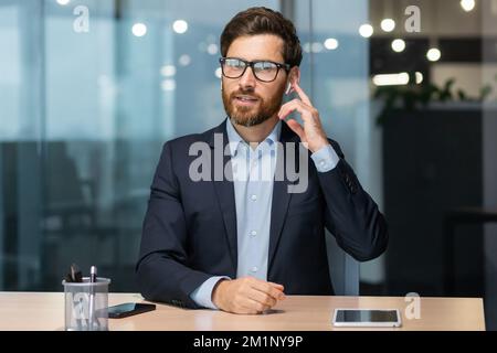 Portrait of a young consultant, businessman. Sitting in the office at the table, talking to the camera, holding headphones, business conference, customer support service. Stock Photo