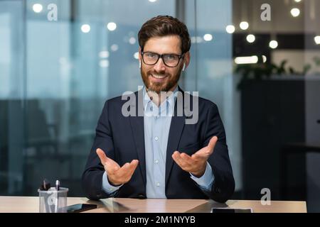 Portrait of a friendly young handsome man sitting in the office at the table wearing headphones and talking to the camera. Business meeting, tutor support, job interview, consultation Stock Photo