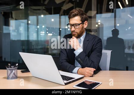 Work in the office of a financial analyst, banker, businessman. A young man sits at the table thoughtfully at a laptop. Stock Photo