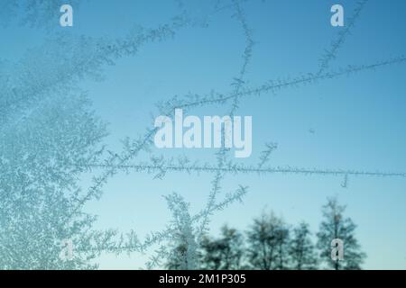 Frost patterns on glass window pane. Frosted glass with blue sky behind. Delicate crystals formed in cold weather. Stock Photo