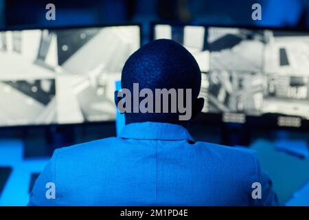 Back view of bald black man looking at surveillance camera footage in dark office, blue and red tones Stock Photo