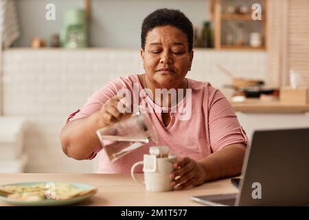 Warm toned portrait of black senior woman making tea during breakfast in cozy kitchen Stock Photo