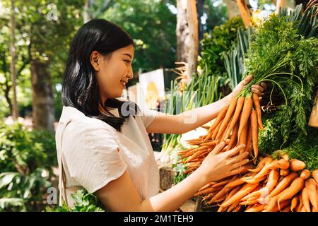 Smiling buyer holding a bunch of carrots. Asian woman choosing vegetables in the local market. Stock Photo