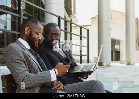 Business meeting of friends outdoors. Two dark-skinned men in suits are sitting on a bench near a city building with a laptop and talking. Partnership Stock Photo