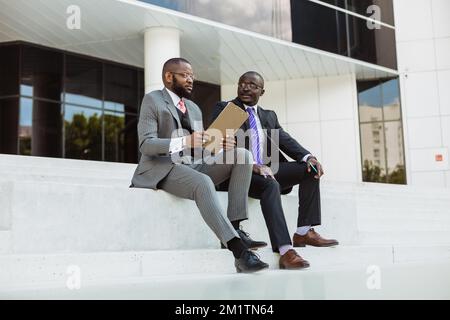 Business outdoor portrait of two dark-skinned friends partners in suits sitting on the concrete steps of the stairs against the backdrop of a city Stock Photo