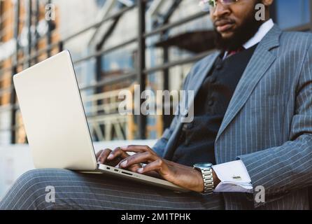 A black businessman in a suit uses a laptop outdoors while sitting on a bench. Close-up Stock Photo