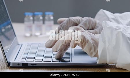 Close-up of covid-19 vaccine stands on wooden table in clinic unrecognizable doctor medical worker scientist in latex gloves typing on laptop makes Stock Photo