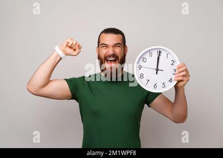 Powerful young man is happy he got all he want in time while holding a clock and making the winner gesture. Stock Photo