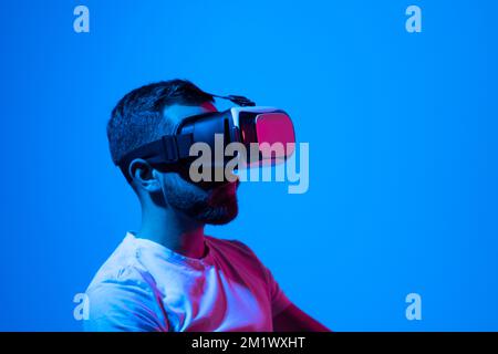 Bearded man in white t-shirt wearing Virtual Reality VR Headset ang playing a game online. VR for gaming. Man play game in VR glasses. Stock Photo