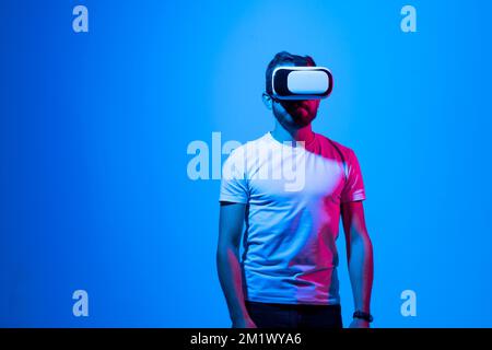 Bearded young man in a white t-shirt wearing virtual reality goggles in modern coworking studio. Stock Photo