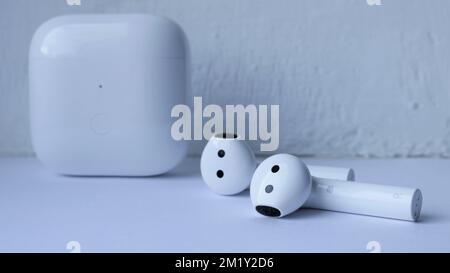 white wireless headphones and a plastic square charging box against a light textured wall, technological still life in shades of white Stock Photo