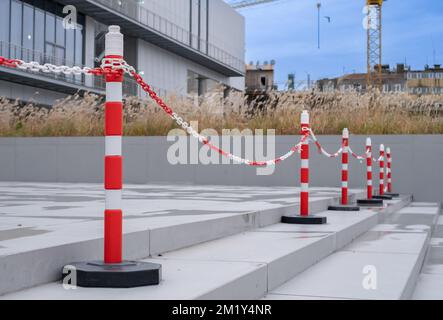 Side view of orange plastic street cones and poles with reflective silver tape near chain link fence at the entrance of a building. Stock Photo