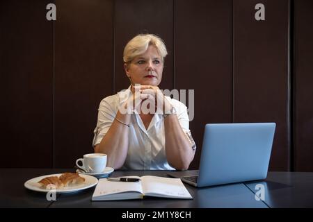 Middle-aged Caucasian woman sitting down with the chin rested on her hands pensively, professional person working in front of an open laptop Stock Photo