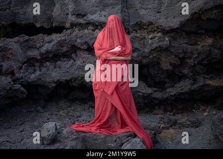 Young woman covered in a long red veil from had to toes looking up with the eyes closed while standing in front of a rocky wall Stock Photo