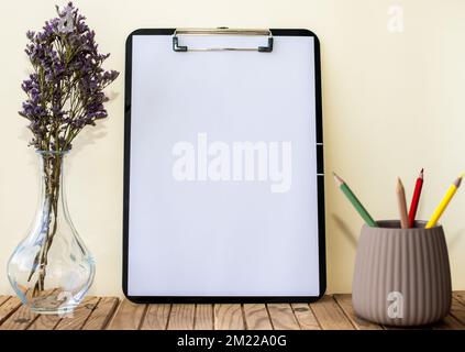 Plain Office Clipboard With Stack Of Pens In A Holder Beside A Flower Vase. Empty Paper Sheet With Pencils Next To Floret Container Placed On Table. Stock Photo