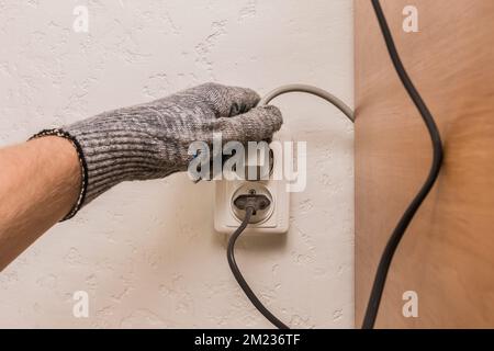 The hand of a construction worker in protective gloves connects a plug to a double socket against the background of a plaster wall. Stock Photo