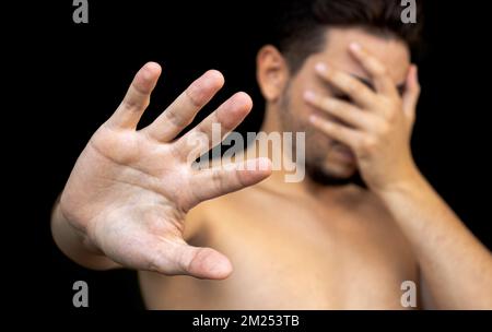 Young sad ashamed man cover his face with his hand to hide on black background suffer from depression Stock Photo