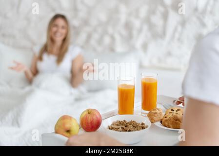 Young man bringing tasty breakfast to bed for his beloved woman, focus on tray with juice, cereals and croissants Stock Photo