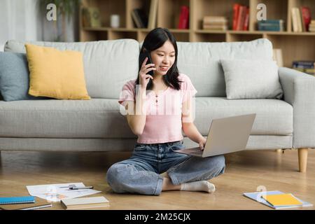 Pretty korean lady having phone conversation, sitting on floor in living room and using laptop, working from home Stock Photo