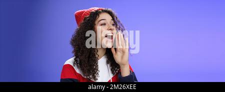 Friend shouting through street to turn attention. Portrait of carefree happy cute woman in beanie and sweatshirt holding palm near opened mouth Stock Photo