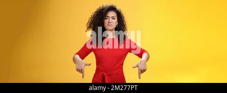 Gloomy disappointed cute silly curly-haired female in red dress frowning upset and making sad smile as pointing down with regret and unsatisfied Stock Photo
