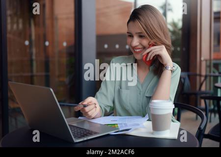 Young business woman sitting at table in coffee shop talking on mobile phone while looking on screen of laptop on table. Stock Photo
