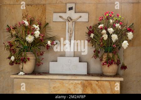 Crucified Jesus Christ figure in altar with withered white and red flowers in marble background inside a Mausoleum cemetery Stock Photo