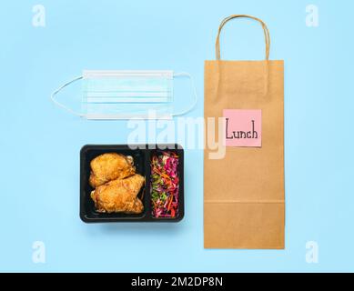 Food box with delicious meal, medical mask, paper bag and sticky note with word LUNCH on color background Stock Photo