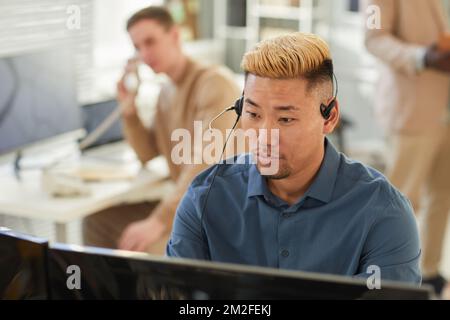 Portrait of young Asian man wearing headset in office and looking at computer screen while working in customer support or surveillance, copy space Stock Photo