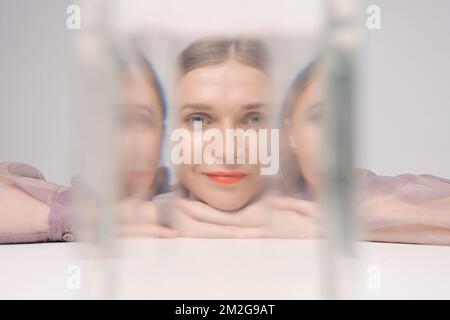 Beautiful woman looking through glass water looks into camera, bright orange lips, on a light background in the studio. Emotional state concept Stock Photo