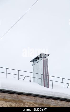 Smoke, rectangular, metal exhaust pipe on the roof of a modern house. The roof of the building is covered with white snow against the gray sky. Cloudy, cold winter day, soft light. Stock Photo