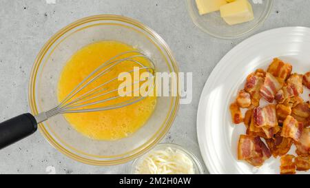 Whipped eggs in a glass bowl, butter and cheese, and crispy fried bacon on a white plate close-up on a light grey background directly from above, Engl Stock Photo