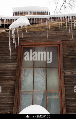 Icy, small icicles hang on the edge of the roof, winter or spring. Plank wall of an old wooden house with windows. Large cascades of icicles in smooth, beautiful rows. Cloudy winter day, soft light. Stock Photo