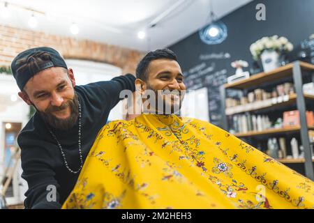 Smiling man at barbershop. Indoor portrait of positive Indian man with yellow patterned cape on his body. Professional caucasian barber and hairstylist. High quality photo Stock Photo