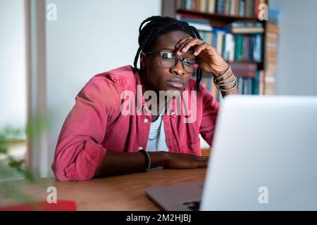 Bored unhappy young African man freelance worker looking at laptop screen cannot concentrate on work Stock Photo