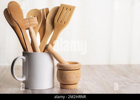 Wooden kitchen utensils such as spoons, forks and tongs inside a jar and next to a kitchen mortar on a wooden counter. Stock Photo