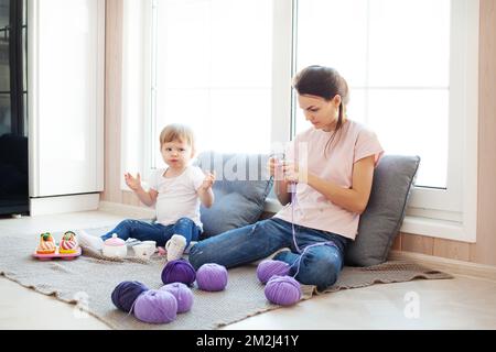 Small kid daughter play while her mother dark hair female knit, doing work or hobby in light wooden interior of house. Family, candid domestic life wi Stock Photo
