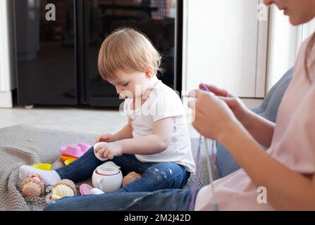 Small child daughter play while her mother knit, doing job or hobby. Family, candid domestic life with baby. Authentic lifestyle Stock Photo