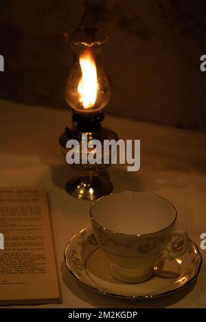 photo of a lit kerazine lamp an empty cup and saucer and an open old book are on the table Stock Photo