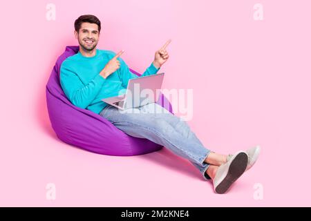 Full size photo of nice young man hold netbook sit beanbag point empty space dressed stylish blue outfit isolated on pink color background Stock Photo
