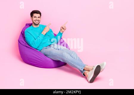 Full length photo of nice young man sitting soft beanbag point empty space dressed stylish blue garment isolated on pink color background Stock Photo